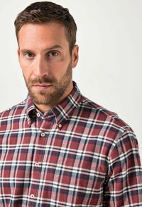 Man met kort bruin haar en baard, gekleed in een rood, wit en blauw geruit overhemd tegen een effen lichte achtergrond.