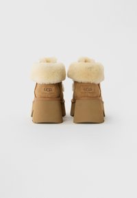 Et par tan kilehælede støvler med tykke cremede pelskraver, set fra bagsiden, med UGG mærkelabels.