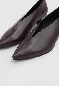 Zapatos de tacón alto de cuero marrón con punta afilada y un diseño con cortes. Textura suave y detalle de costura mínima, sobre un fondo claro.