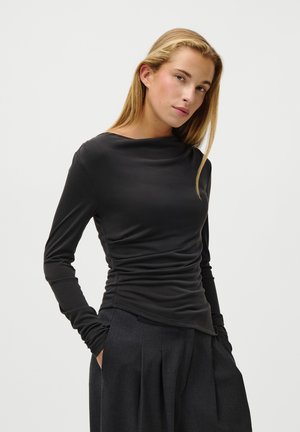 Haut noir à manches longues avec un col bateau et une texture froncée, associé à un pantalon sombre plissé. Tissu lisse, design minimaliste.