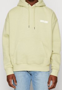Světle zelená mikina s kapucí, přední kapsou, stahovací šňůrkou a bílou nášivkou s logem Calvin Klein na horní části levého hrudníku. Měkká bavlněná tkanina.