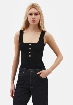 Blondynka ubrana w czarny top bez rękawów z czterema złotymi ozdobnymi guzikami i ciemne jeansy z wysokim stanem, ręka w kieszeni, neutralny wyraz twarzy.