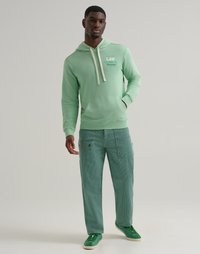 Uomo che indossa una felpa leggera verde con lacci bianchi, pantaloni cargo a strisce turchesi e sneakers verdi, in piedi su uno sfondo semplice.