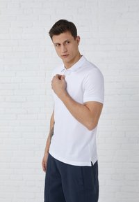 Witte polo met korte mouwen en een klassieke kraag, met een knoopsluiting met twee knopen en zijsplitten, gedragen met donkerblauwe shorts.