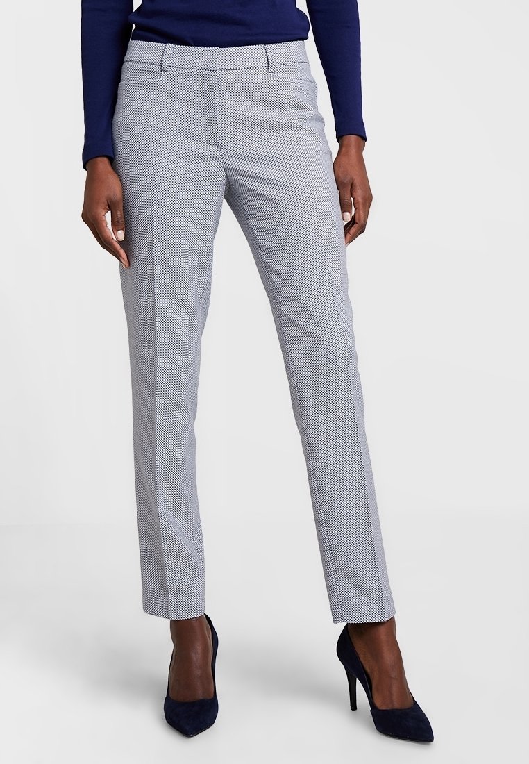Femme portant un pantalon gris ajusté à motifs et des chaussures à talons hauts bleu marine, debout devant un fond uni.