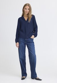 Blusa blu navy a maniche lunghe con scollatura a V, abbinata a jeans blu a taglio comodo. Le scarpe sono ballerine nere dal design elegante e minimal.