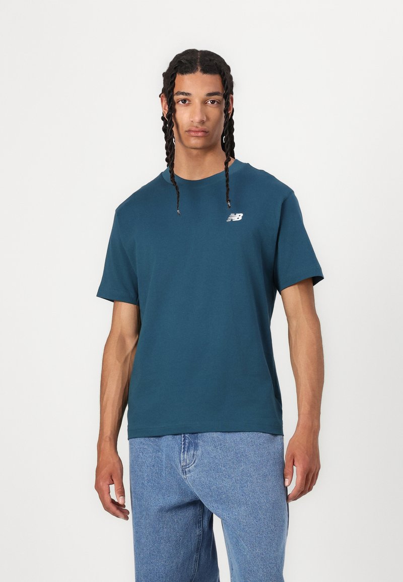 Teal bomull T-skjorte med korte ermer, rund hals og en liten hvit logo på venstre bryst; kombinert med lyseblå jeans.