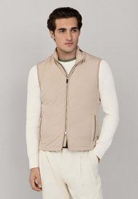 Jeune homme portant une veste matelassée beige sur un pull côtelé blanc avec un pantalon plissé crème, debout avec une main dans la poche.