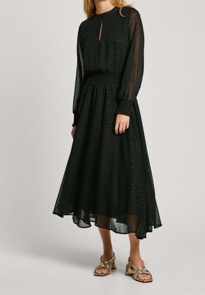 Femme portant une robe midi noire à manches longues en tissu transparent à pois avec une taille smockée, associée à des sandales à talons carrés dorées à brides.