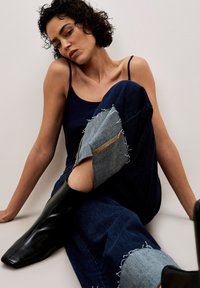 Ein marineblaues Tanktop kombiniert mit dunkelblauer Jeans, die mit Denim-Patches, abgenutzten Details und einem sichtbaren Reißverschluss versehen sind. Schwarze Stiefeletten runden das Outfit ab.