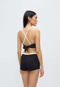 Σετ μαγιό σε μαύρο χρώμα με ριμπ υφή, λουράκια που διασταυρώνονται στην πλάτη και φιόγκο στη μέση της πλάτης. Διακοσμημένο με μια κρεμ-χρωματιστή ταινία.