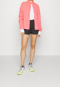 Osoba ubrana w koralową bluzę na zamek, białą koszulę, czarne szorty sportowe i białe trampki z neonowo-żółtymi podeszwami, stojąca na jednolitym tle.