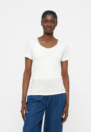 Femme à la peau foncée et aux cheveux tirés en arrière, portant un haut blanc à manches courtes avec des détails en dentelle et un pantalon ample bleu, posant devant un fond uni.