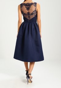 Ärmlös marinblå midi-klänning med plisserad kjol och genomskinlig spetsbak, kombinerad med grå sammet högklackade ankelremssandaler.