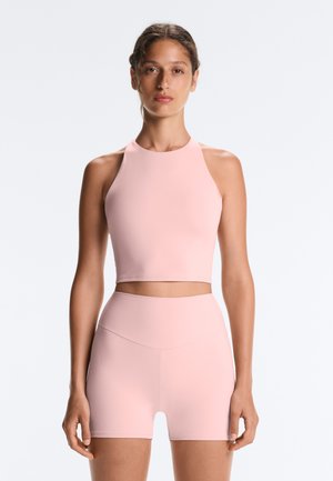 Kvinde iført en lyserød ærmeløs crop top og matchende højtaljede shorts står foran en lys, ensfarvet baggrund.