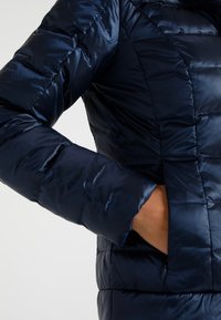 Laivastonsininen toppatakki kiiltävällä pinnalla, tikattu muotoilu ja sivutaskut. Siinä on vartalonmyötäinen siluetti ja tikatut yksityiskohdat.