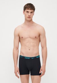 Svarta bomullskalsonger med resårmidja som pryds av en blå Nike-logotyp. Modellen har kort brunt hår och ljus hudton.