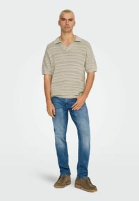 Beige gebreide polo met korte mouwen, voorzien van een gestructureerd patroon, gecombineerd met lichtblauwe jeans en bruine suède veterschoenen.