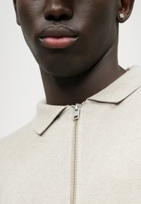 Beige strikpolo med lynlås, der har en flad krave og en subtil struktur, suppleret med sølvhardwaredetaljer.