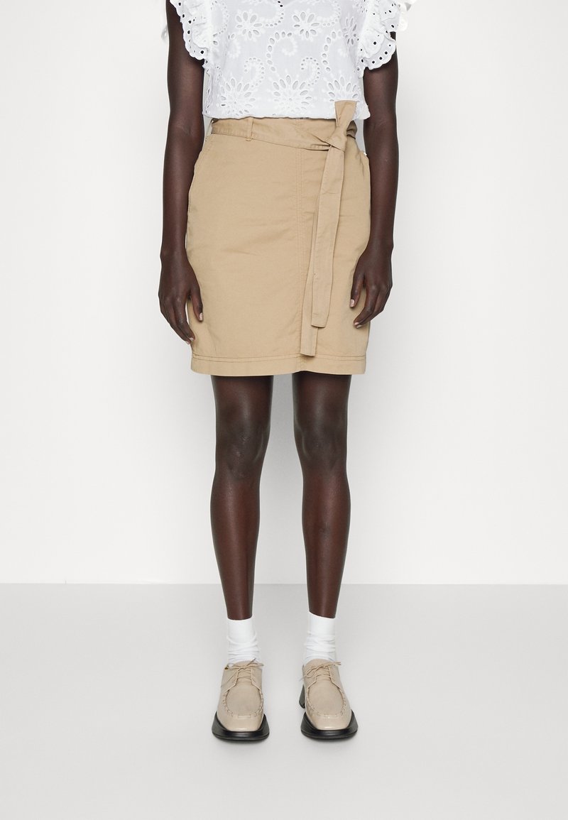 Personne portant une jupe portefeuille beige avec lien, un haut blanc à œillets floraux, des chaussettes blanches et des chaussures beige à lacets, debout devant un fond uni.