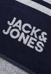 Serviette en coton bleu marine avec une surface douce et texturée, ornée du logo blanc "JACK & JONES" et de deux bandes blanches horizontales.