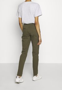 Pantalon olive vert ajusté avec une coupe slim, deux poches arrière, et des passants pour ceinture, associé à un t-shirt manches courtes gris clair et des baskets blanches.
