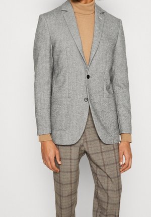 Man draagt een lichtgrijs visgraatblazer, een camelkleurige coltrui en beige geruite broek, staand tegen een effen witte achtergrond.