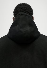 Črna kapuča s kapuco, ki ima gosto kapo, teksturirano blago in vidno šivanje na vratu in ramenskih šivih, prikazana s hrbta.