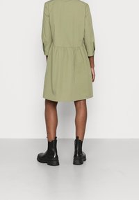 Robe vert olive avec une silhouette décontractée, des manches trois-quarts, une taille froncée et un ourlet droit. Assortie à des bottines noires à chevilles.