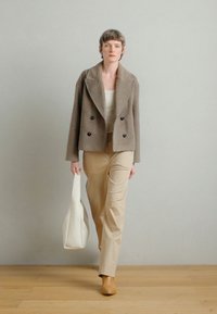 Pruun kahepoolse nööpidega jakk, beežid kitsad püksid, pruunid kingad ja valge kandekott; iseloomustavad pehmed tekstuurid ja istuv lõige.