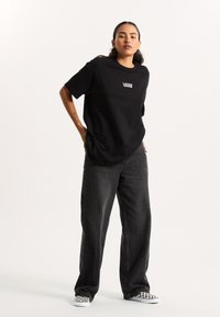Black oversized t-shirt with Vans logo, paired with loose black jeans and black-and-white checkerboard slip-on shoes. Smooth cotton texture.