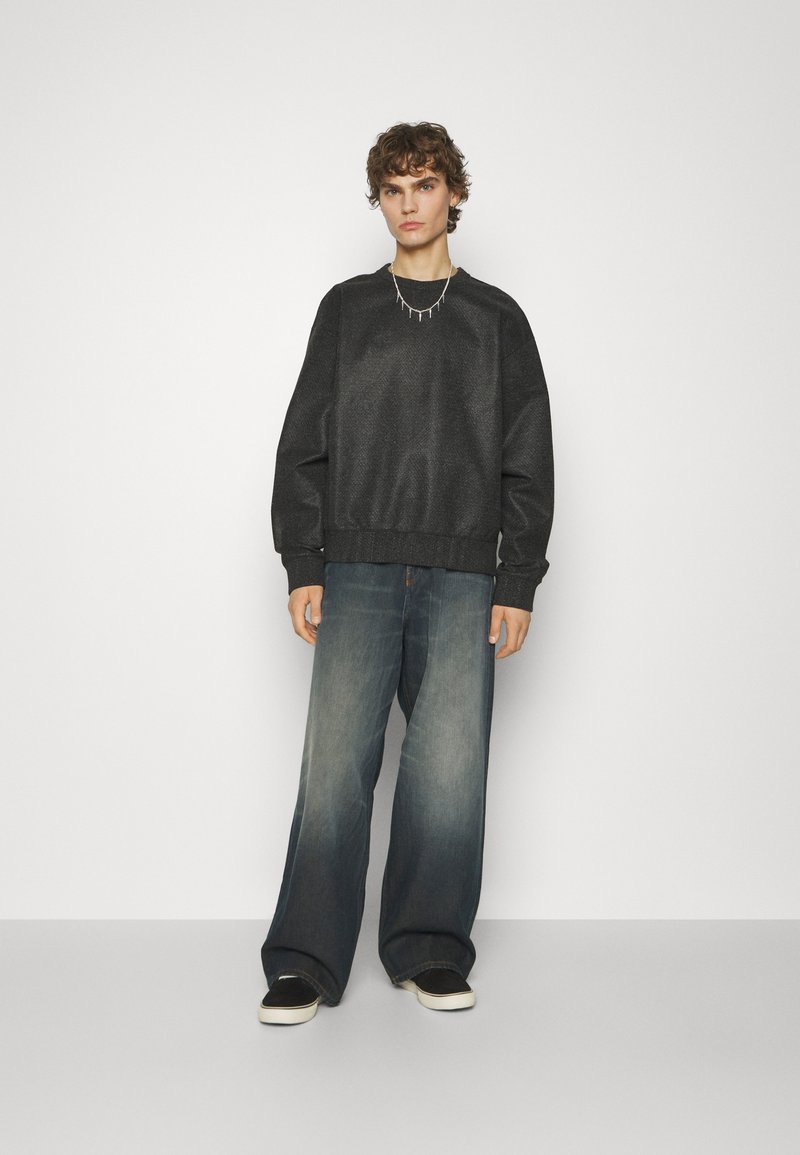 Czarna, fakturowana bluza z ribbed hem, w połączeniu z szerokimi, wyblakłymi niebieskimi dżinsami i czarnymi butami wsuwanymi. Prosty srebrny naszyjnik jako detal.