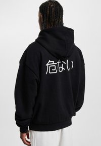 Un sweat à capuche noir avec une poche kangourou, de coupe surdimensionnée, et un design en texte blanc au dos. Matériau doux et épais avec des poignets côtelés.