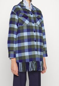 Skjortejakke i uldblanding med tern i blå, grøn og marineblå, to forlommer, knaplukning og frynser ved kanten.