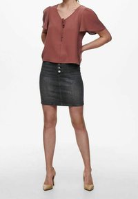 Blouse rose à manches courtes avec détail noué, associée à une jupe en jean longueur genou avec boutons sur le devant et coupe ajustée. Talons beiges.