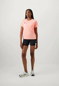 Žena stoji u breskvastom majici kratkih rukava, crnim šortsevima i bijelim tenisicama za trčanje, naspram jednostavne svijetle pozadine.