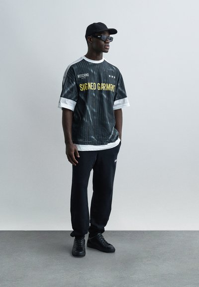 Άνδρας που φοράει μαύρο καπέλο, γυαλιά ηλίου, μαύρη και λευκή μπλούζα Moschino "Signed Garment", μαύρο παντελόνι και μαύρα αθλητικά παπούτσια, στέκεται μπροστά σε απλό φόντο.