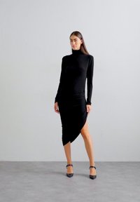 Rochie neagră cu mânecă lungă, având un design confortabil, o linie asimetrică și o textură moale, asociată cu pantofi cu toc înalt negri.