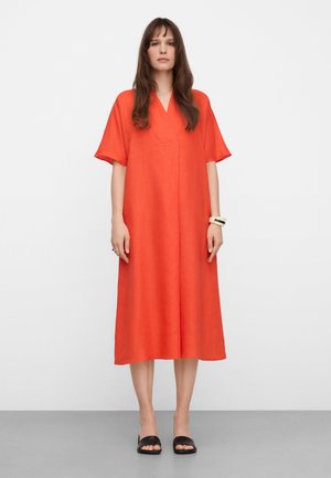 Frau mit langen braunen Haaren, die ein locker sitzendes, knieumspielendes oranges Kleid mit kurzen Ärmeln, schwarze Sandalen mit offenen Zehen und ein weißes Armband trägt.