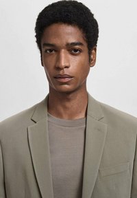 Light gray blazer with a textured finish, single button closure, and a pocket, worn over a beige crew-neck t-shirt. Neutral tones.
