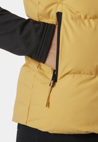 Gelbe Puffy-Weste mit schwarzem Langarmshirt darunter. Verfügt über eine Reißverschlusstasche mit kontrastierendem Reißverschluss und Ziehschlaufe. Texturierter Stoff.