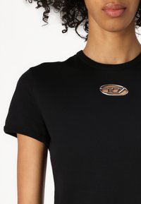 Svart bomull T-skjorte med korte ermer, med et sølvfarget ovalt logoakssent ved brystet. Jevn tekstur og tettsittende design.