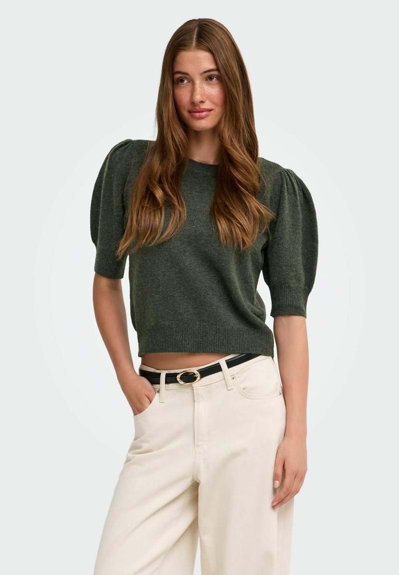Femme aux longs cheveux bruns portant un haut vert foncé à manches bouffantes et un pantalon taille haute écru avec une ceinture noire sur un fond neutre.