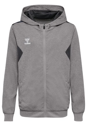 Garçon debout, les mains dans les poches, portant un sweat à capuche gris zippé, un short de sport noir, des chaussettes blanches et des baskets noires sur fond uni.