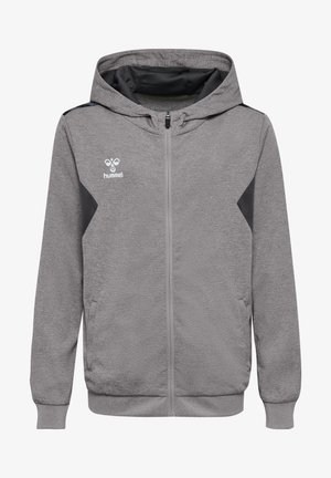 Junge steht mit den Händen in den Taschen, trägt einen grauen Kapuzenpullover mit Reißverschluss, schwarze Sportshorts, weiße Socken und schwarze Sneakers vor einfarbigem Hintergrund.