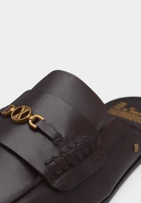 Mocassin en cuir marron foncé avec détail en maillon métallique doré et surpiqûres visibles sur la tige.