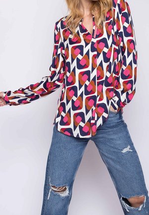 Vrouw draagt een blouse met lange mouwen met een geometrisch patroon van roze, rood en marineblauwe vormen en gescheurde blauwe spijkerbroek, poseert met één arm omhoog.