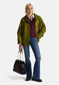 Grüne wasserdichte Jacke mit großem Kragen, bordeauxroter Pullover, blaue Schlagjeans und eine schwarze Handtasche mit goldenen Kettenriemen.