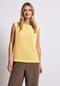Top giallo senza maniche con superficie texturizzata e scollo rotondo, abbinato a pantaloni in lino marroni, creando un outfit casual.