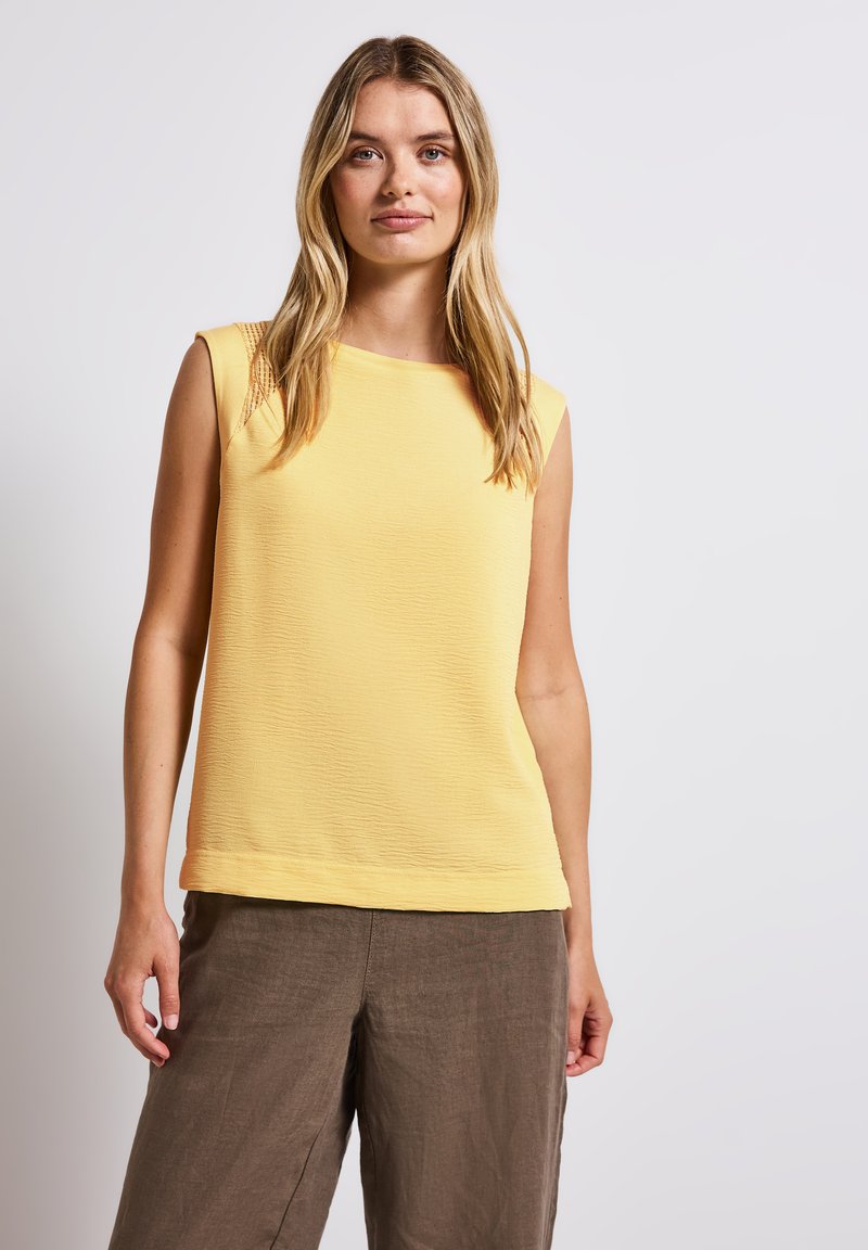 Top giallo senza maniche con superficie texturizzata e scollo rotondo, abbinato a pantaloni in lino marroni, creando un outfit casual.